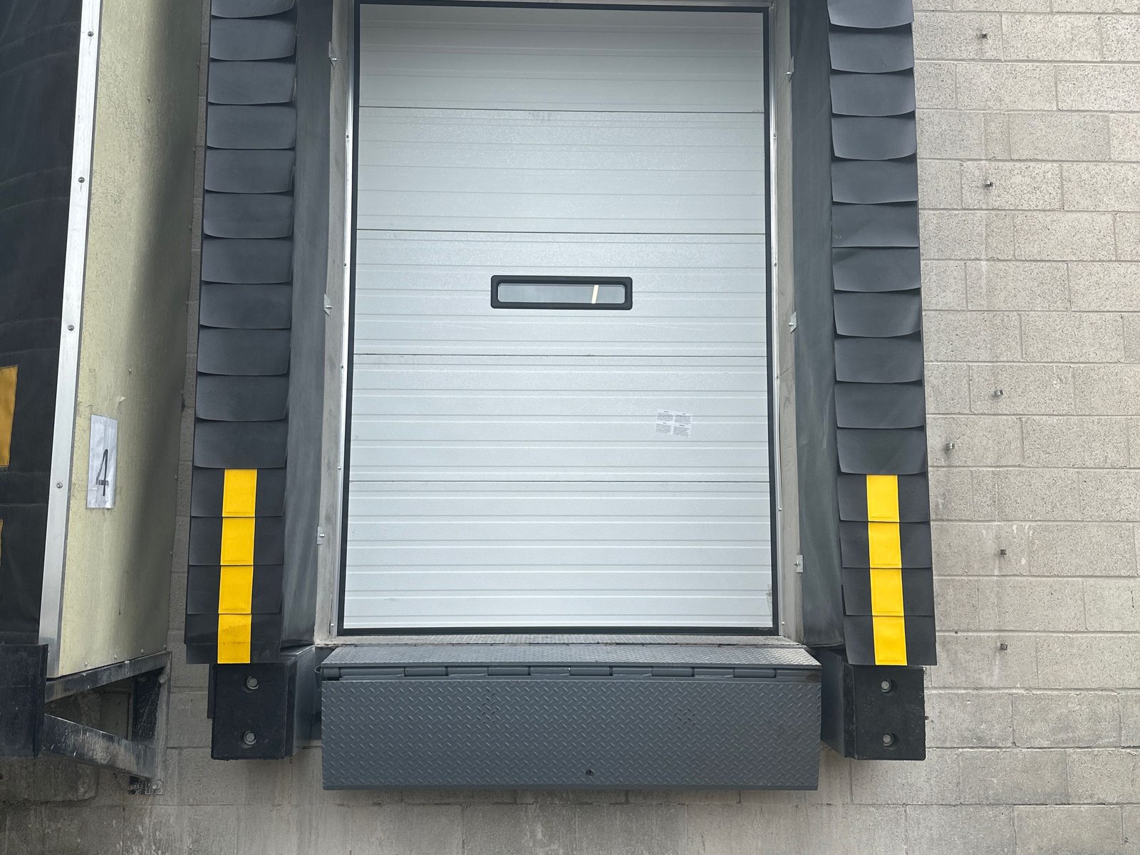 A loading dock position with a black compression dock seal, yellow-striped jamb pads, laminated rubber bumpers and a diamond-plate edge-of-dock leveler under a white sectional door, installed by Canuck Door Systems