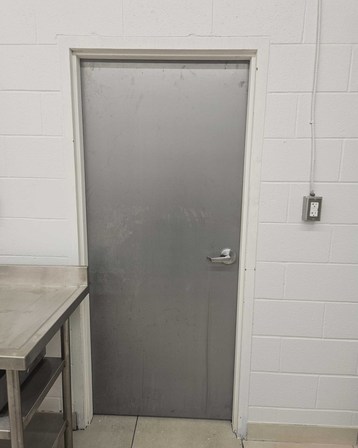 A black hollow metal door under a surface-mounted automatic operator, with a push-to-open actuator and a push-to-lock switch on the wall beside it, installed by Canuck Door Systems