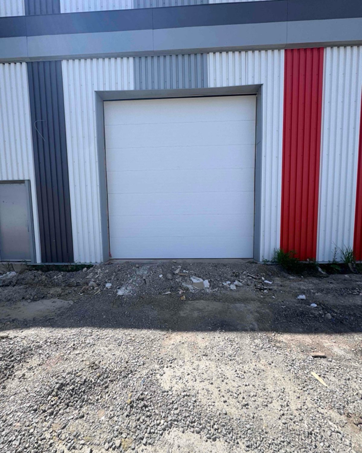 A newly installed white sectional overhead door in a steel-clad industrial unit, with the ground outside still unfinished, installed by Canuck Door Systems
