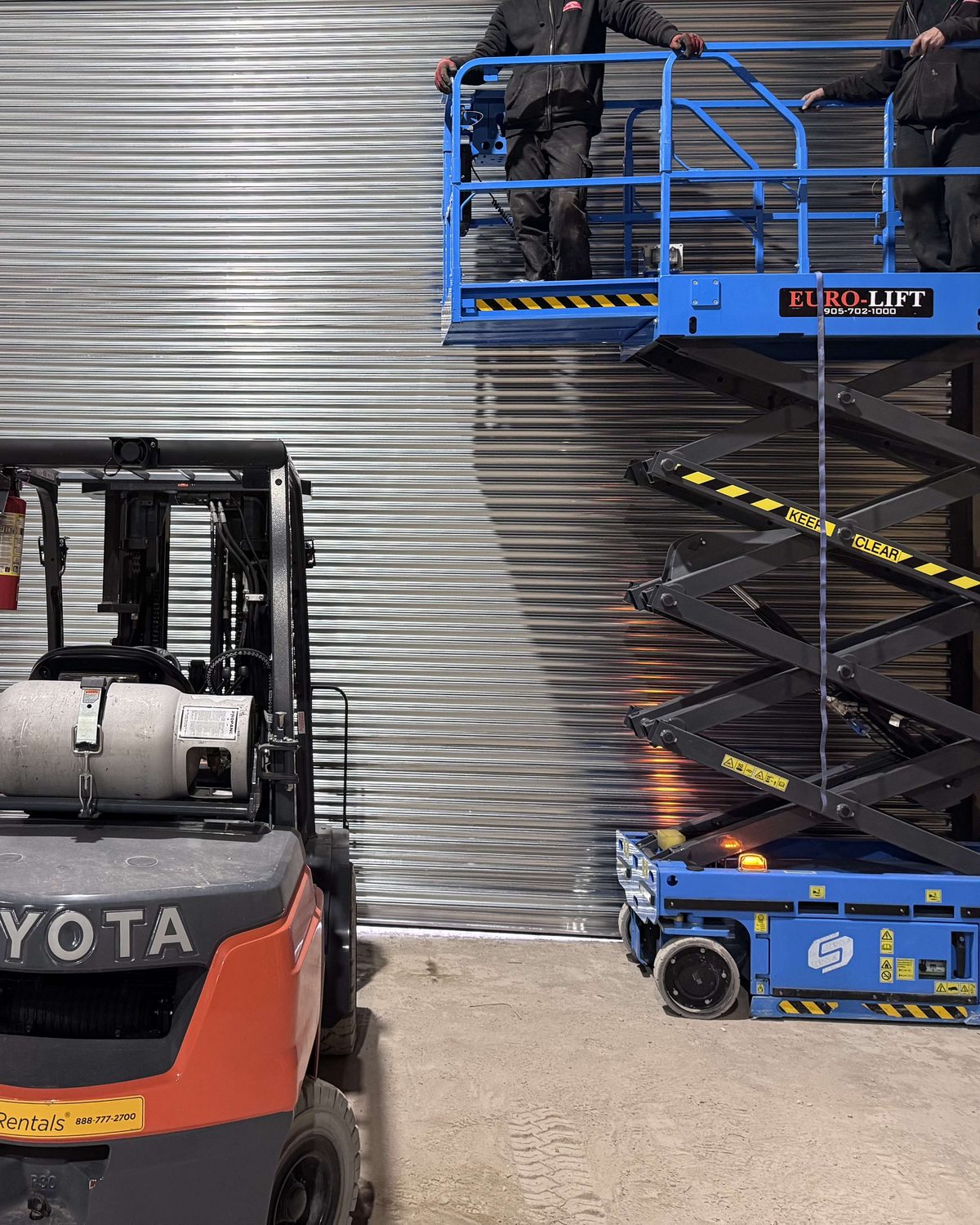 Two Canuck Door Systems technicians on a raised scissor lift working on a rolling steel curtain inside an industrial building, with a forklift and a second lift on the floor below