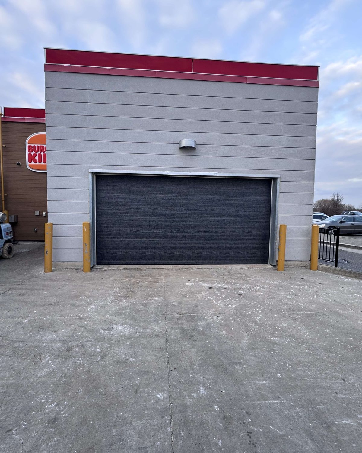 A dark grey insulated roll-up overhead door in a precast service building with yellow bollards either side, installed by Canuck Door Systems