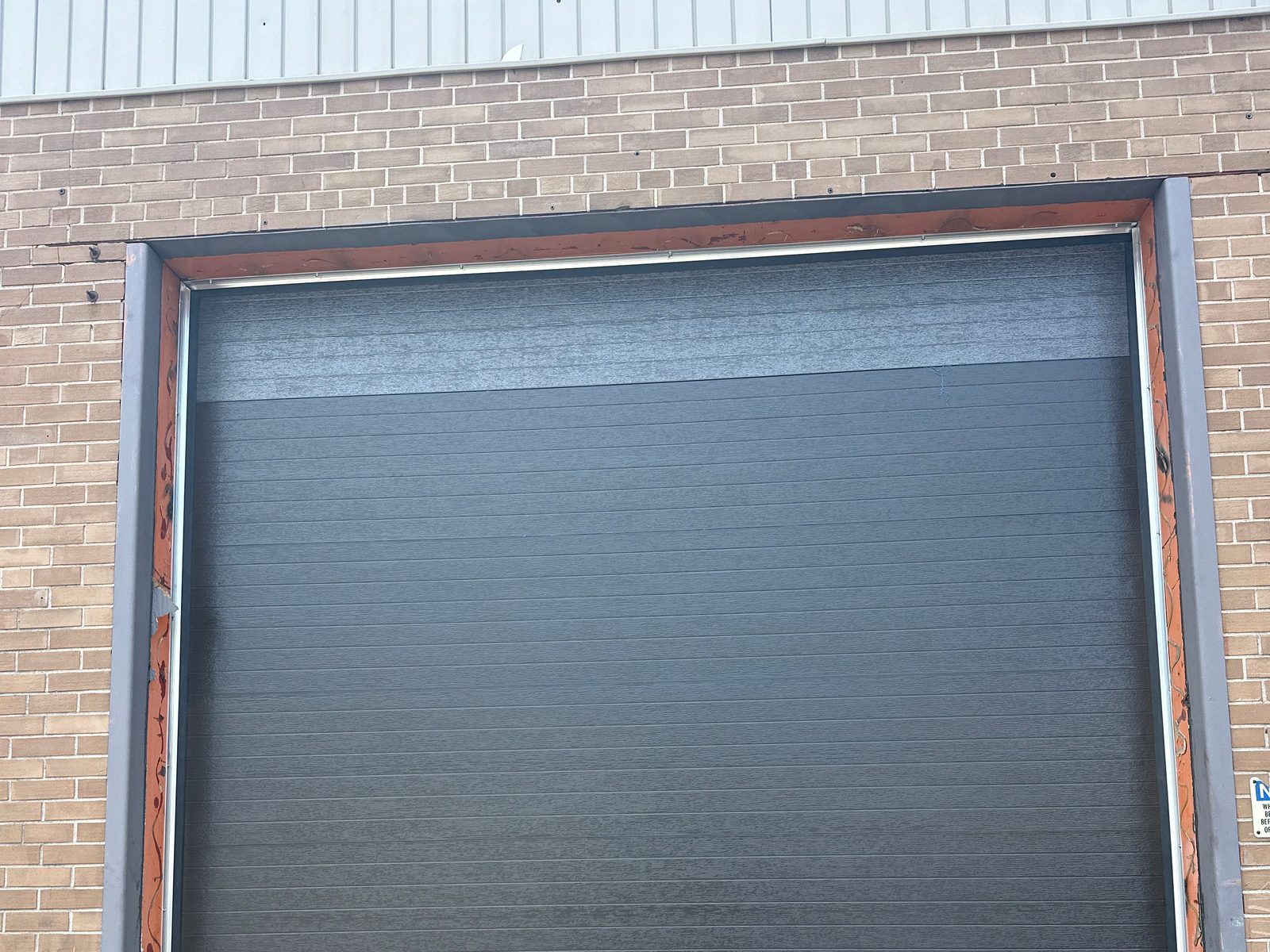 A charcoal sectional overhead door closed in a brick opening with steel jamb guards, on a Toronto industrial unit, installed by Canuck Door Systems