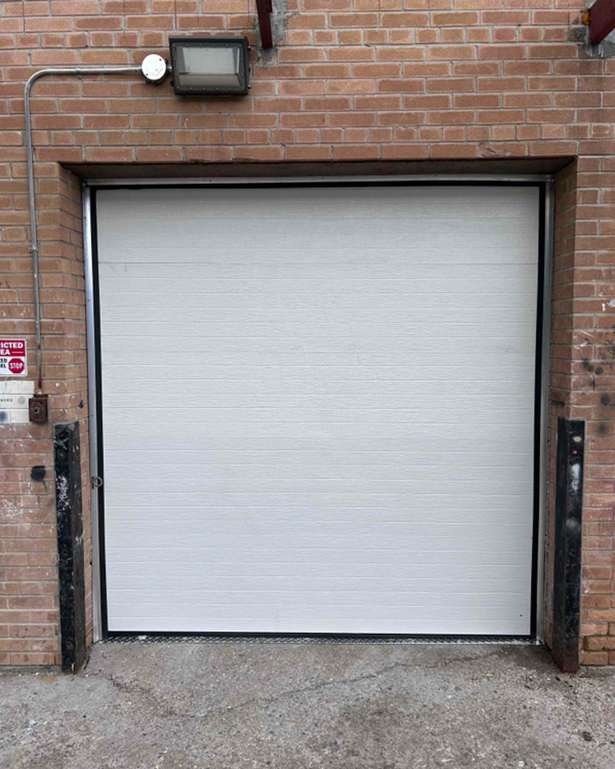 A white sectional overhead door closed in a brick opening protected by steel jamb guards, installed by Canuck Door Systems