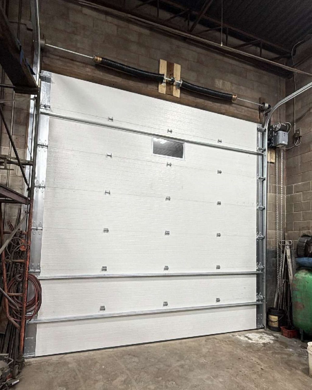 A torsion spring on its shaft mounted on the header above a white sectional overhead door, with the cable drums, track and a jackshaft operator beside the opening, serviced by Canuck Door Systems