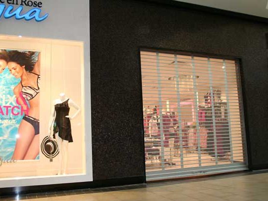 A perforated steel rolling security grille closed across a lit mall shopfront, with the merchandise visible through the curtain, installed by Canuck Door Systems
