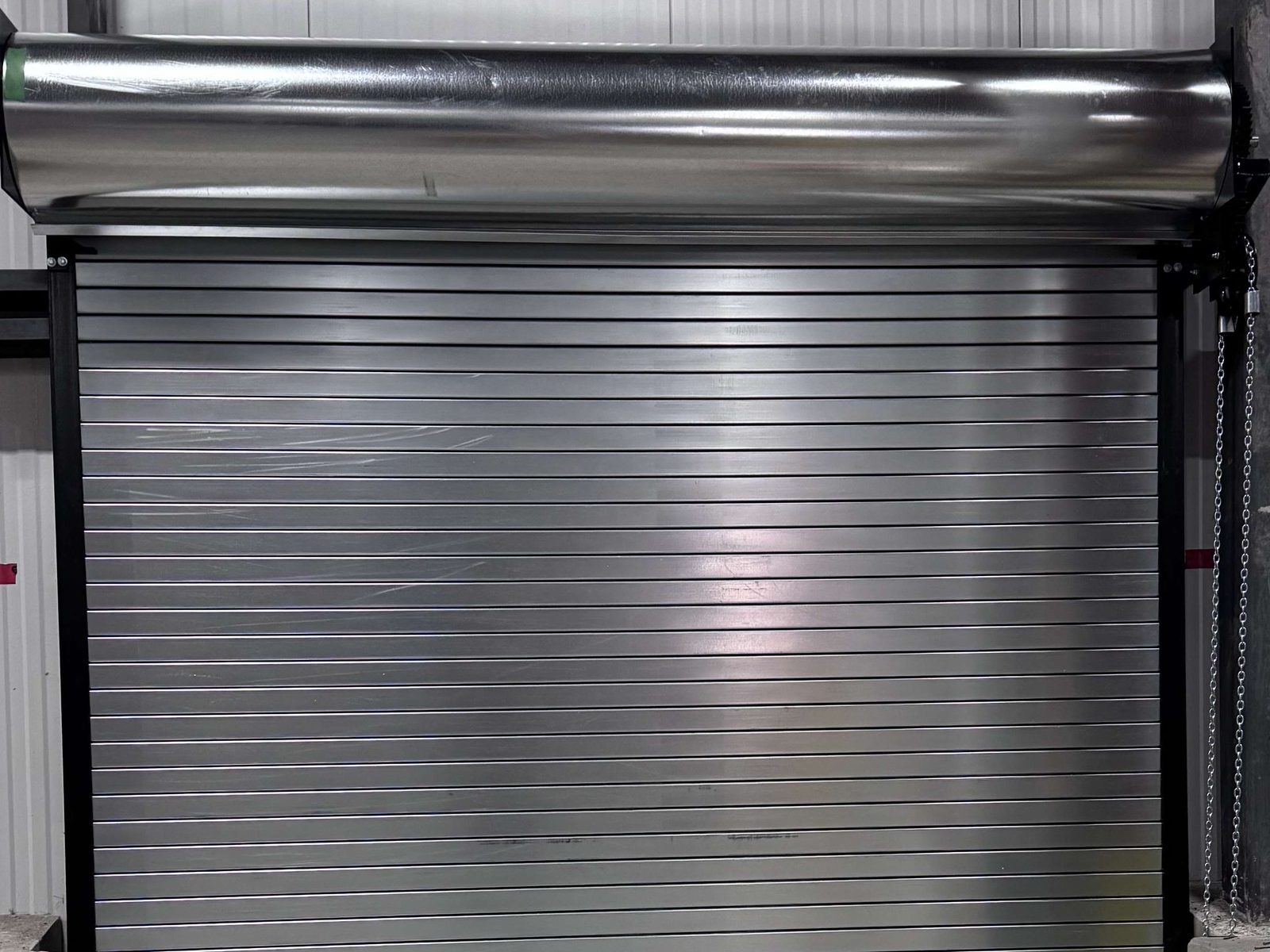 A galvanized roll-up door closed in an industrial opening, with the coil and barrel mounted on brackets above the head and the hand chain hanging beside it, installed by Canuck Door Systems