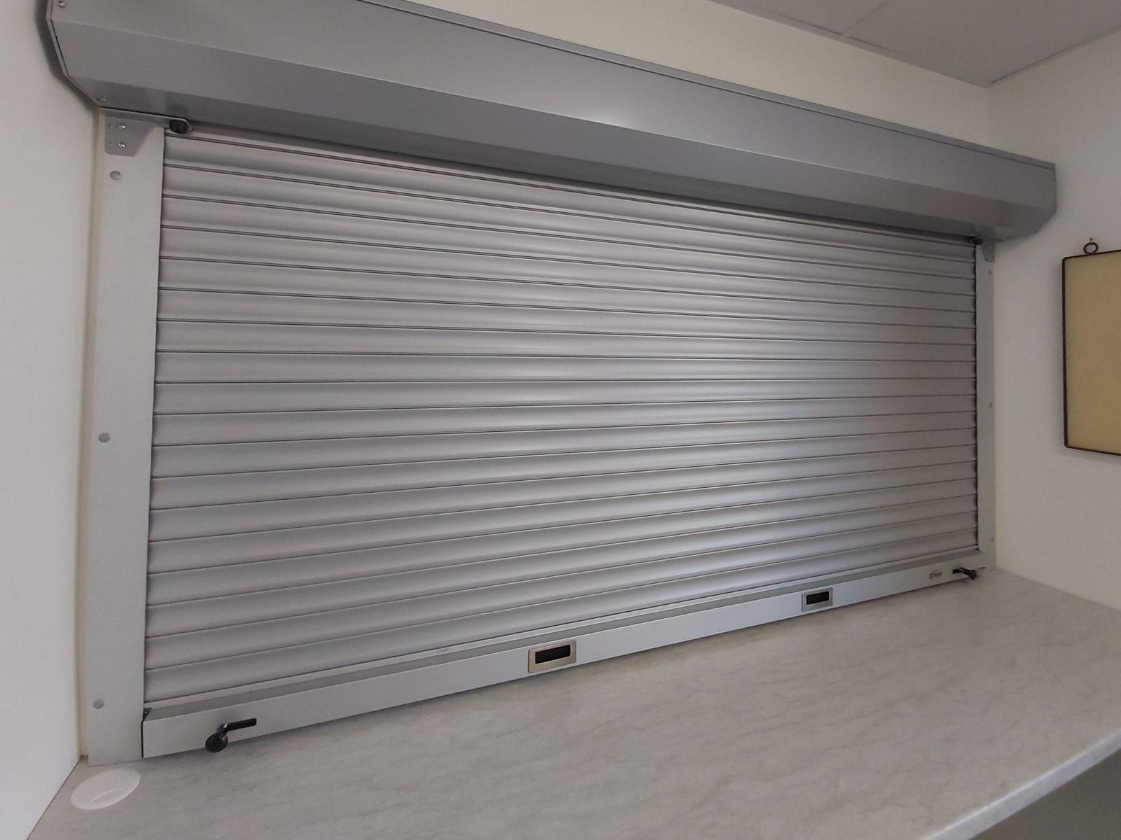 A silver aluminum rolling counter shutter closed down onto a servery counter, with its hood mounted above the opening, installed by Canuck Door Systems
