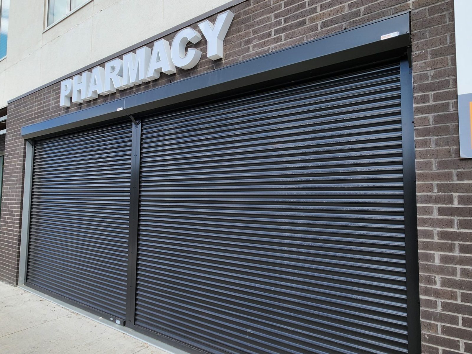 Two black rolling security shutters closed across a pharmacy storefront, with the head boxes and side guides fixed to the brick, installed by Canuck Door Systems
