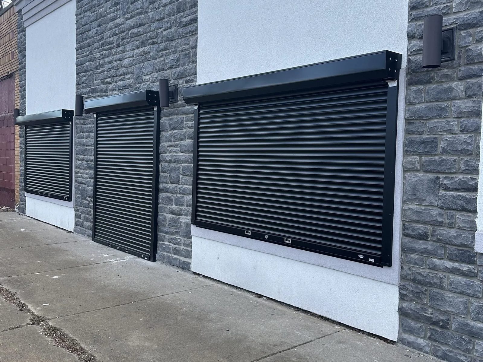 Three black rolling security shutters closed across the window openings of a stone-clad building, with surface-mounted hoods and side guides, installed by Canuck Door Systems
