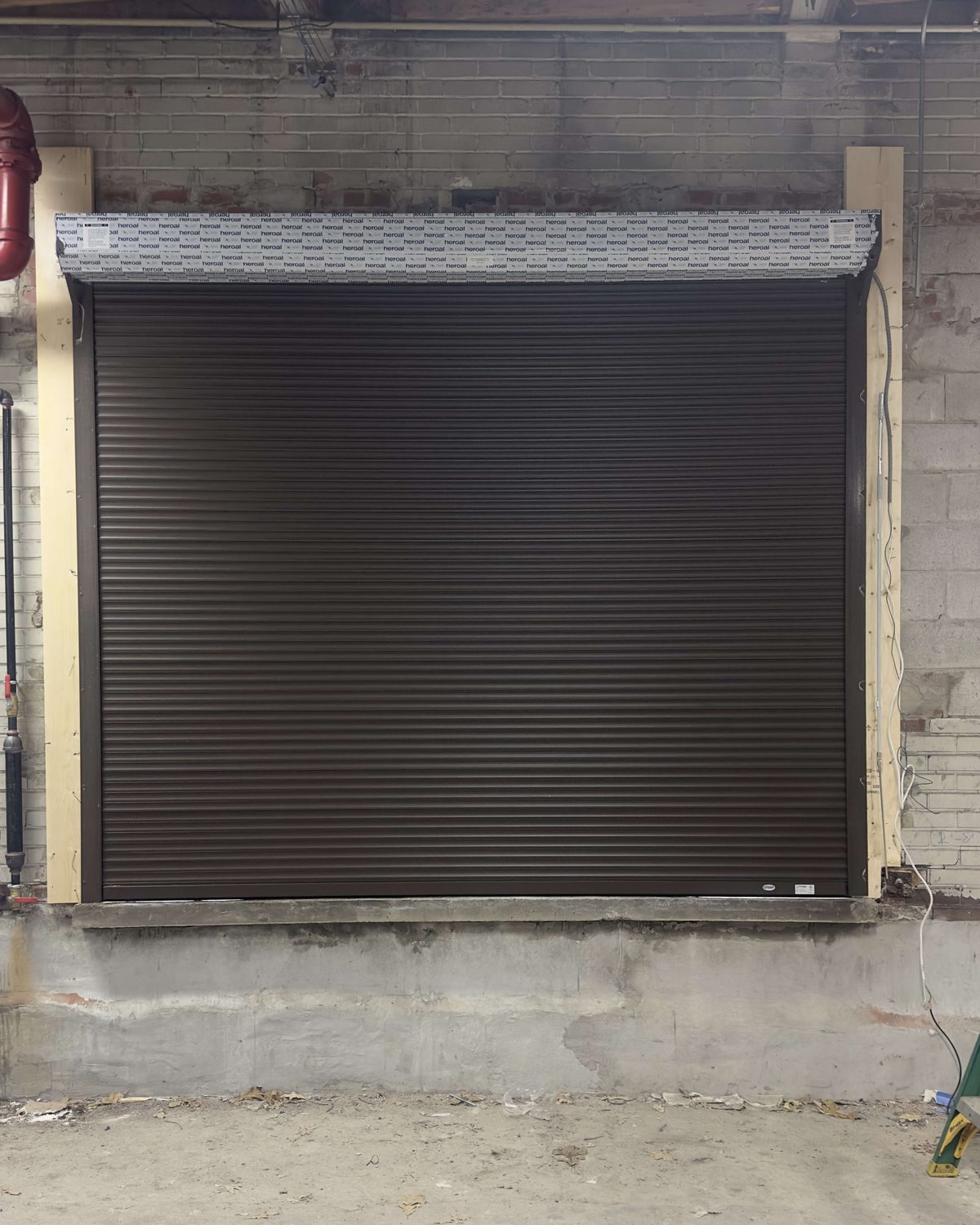 A brown slat rolling shutter closed in a brick opening, with the coil hood fitted above the head, installed by Canuck Door Systems