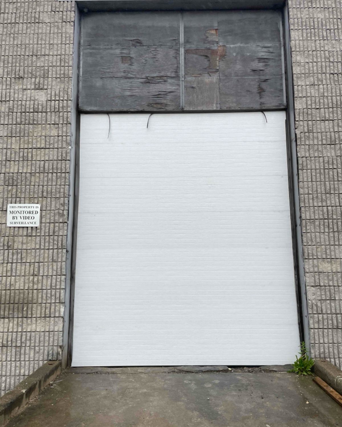 A white sectional overhead door filling a tall block-wall opening, with the original infill panel above it left in place, installed by Canuck Door Systems