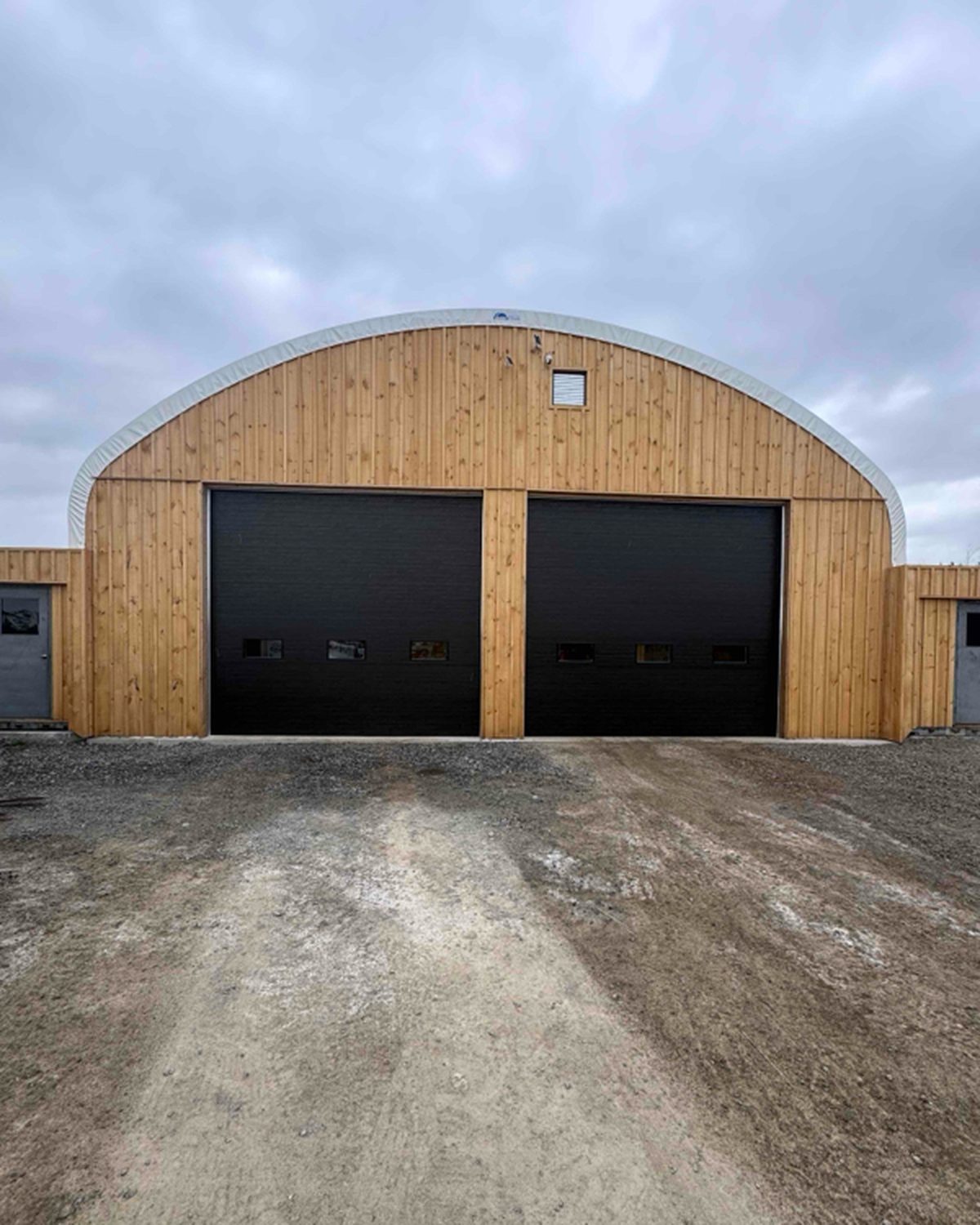 A pair of black sectional overhead doors with vision panels, set into the timber-clad end wall of an arched industrial building, installed by Canuck Door Systems