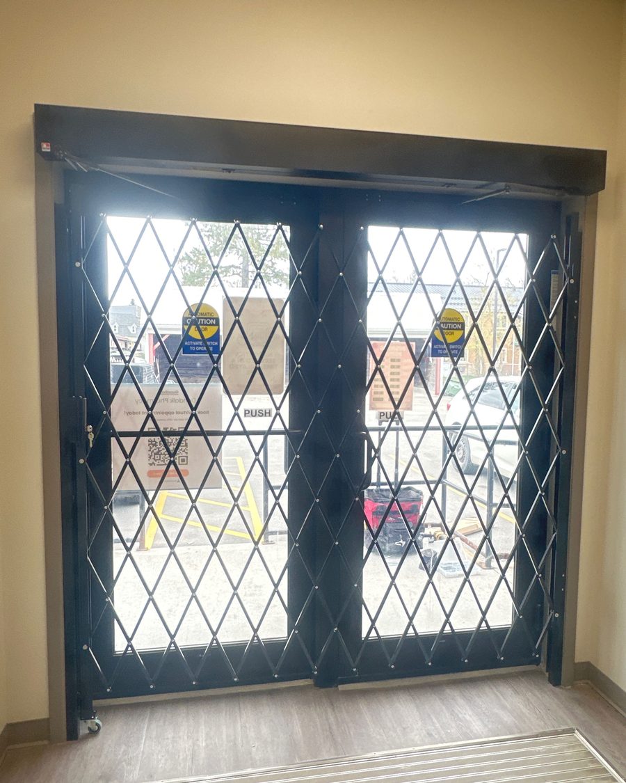A pair of glazed storefront doors closed off by a diamond-pattern folding security grille inside a pharmacy, installed by Canuck Door Systems