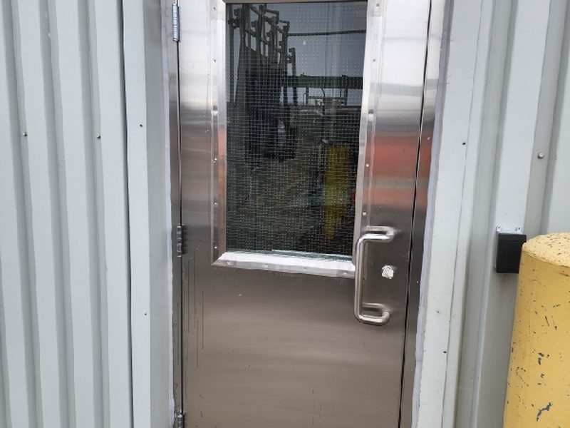 A stainless steel door with a wire-glass vision lite and a stainless pull, set into a metal-clad plant wall, installed by Canuck Door Systems