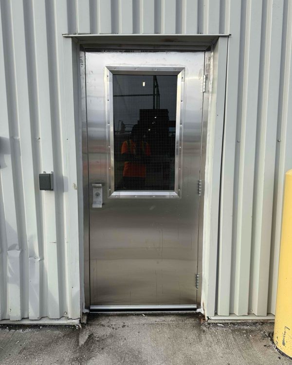 A stainless steel door with a vision lite in a metal-clad industrial wall, installed by Canuck Door Systems