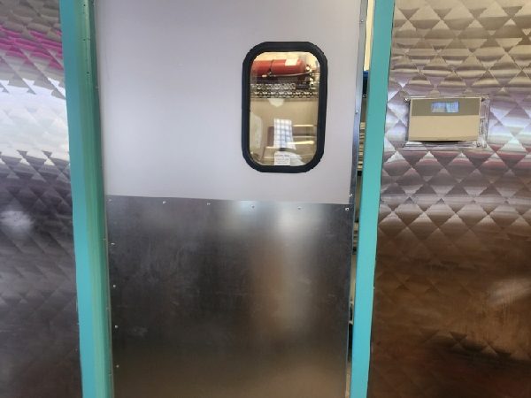 A double-acting traffic door with a vision window and stainless steel kick plate, set in a teal-painted frame in a commercial kitchen, installed by Canuck Door Systems
