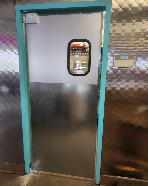A commercial kitchen traffic door with a rounded vision window and brushed stainless lower panel, installed by Canuck Door Systems
