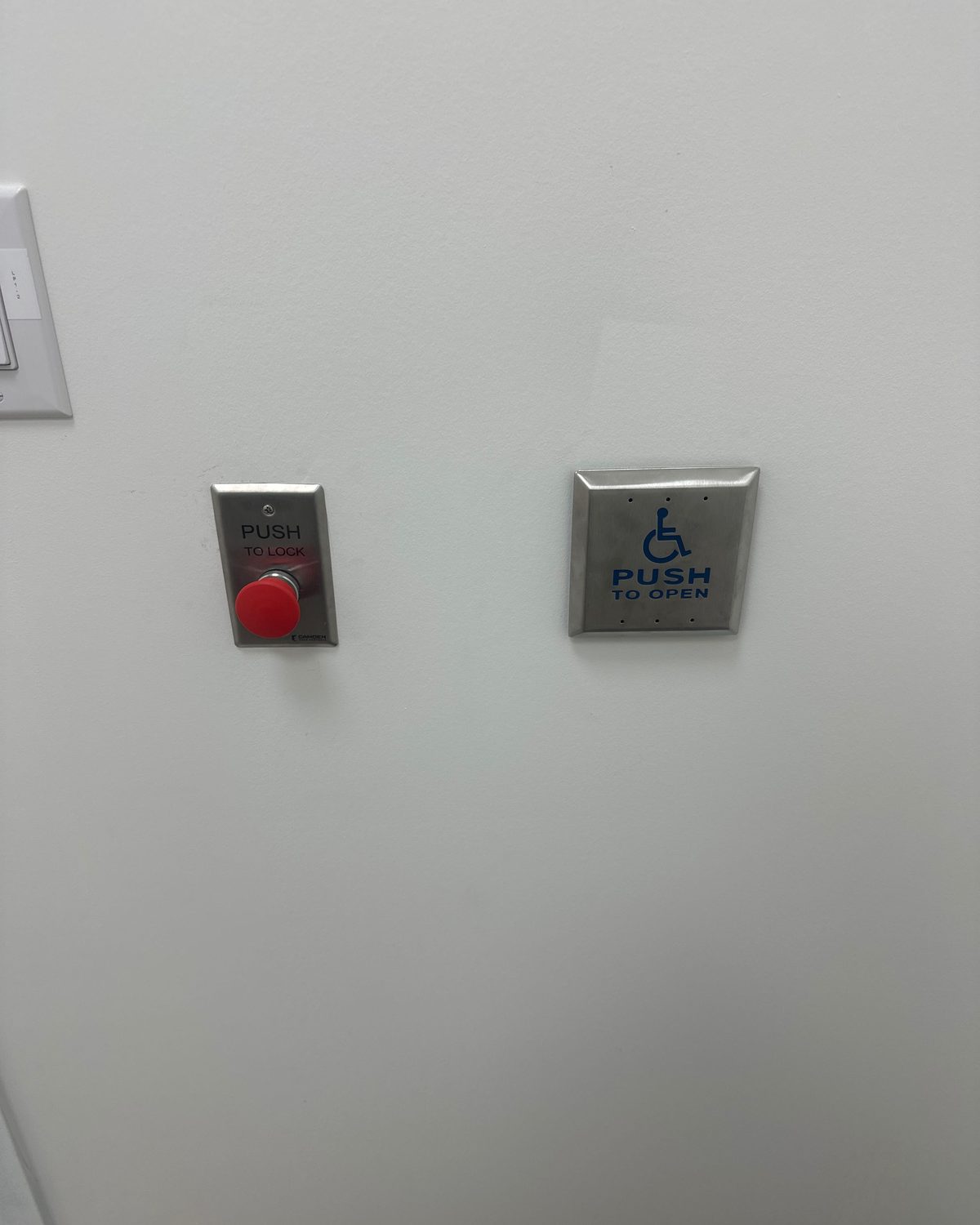 A stainless push-to-open actuator plate beside a red emergency call button on a universal washroom wall, installed by Canuck Door Systems