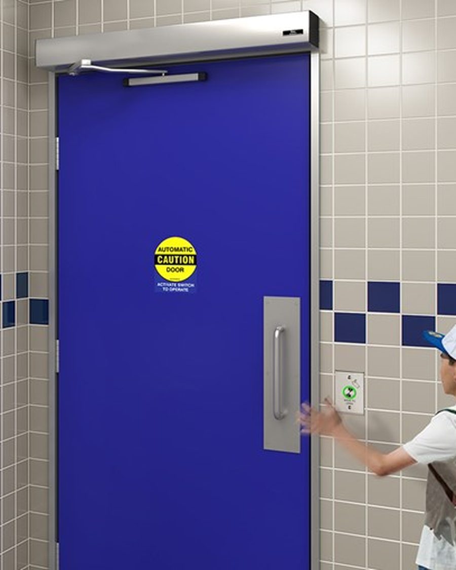 A universal washroom door with an operator in the header and an automatic caution decal, being opened by someone pressing the wall-mounted actuator plate beside the opening
