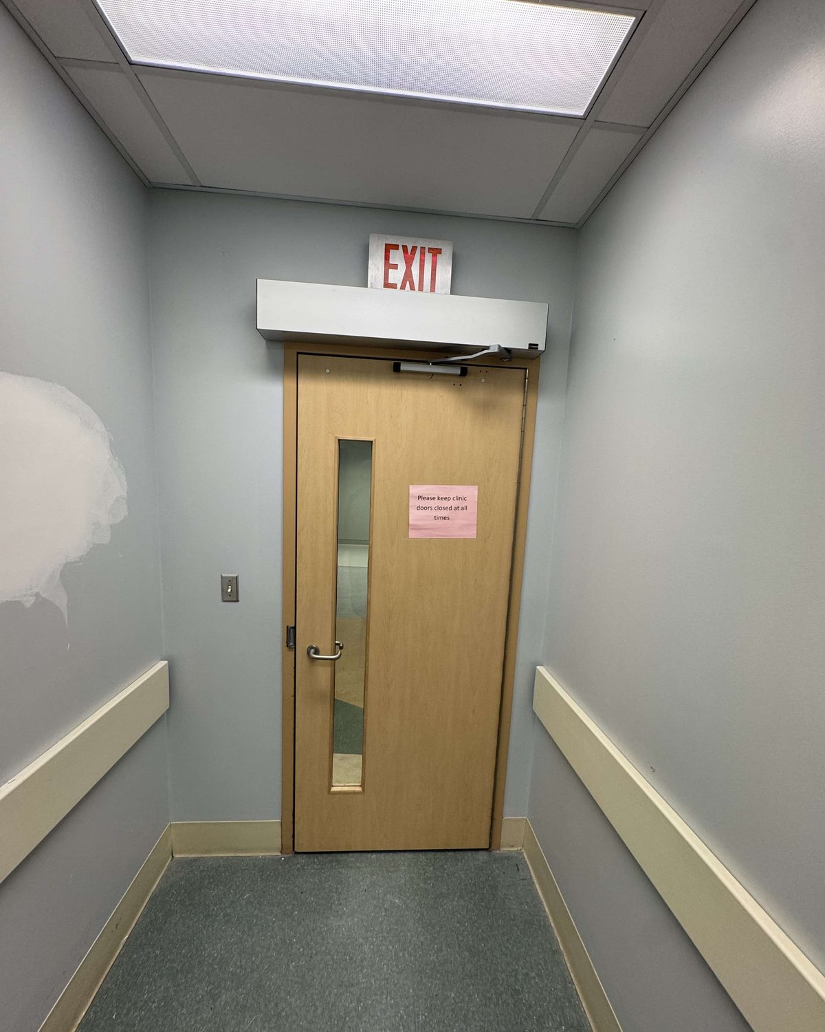 A wood-veneer clinic corridor door with a narrow vision lite and an overhead closer, beneath an exit sign, installed by Canuck Door Systems
