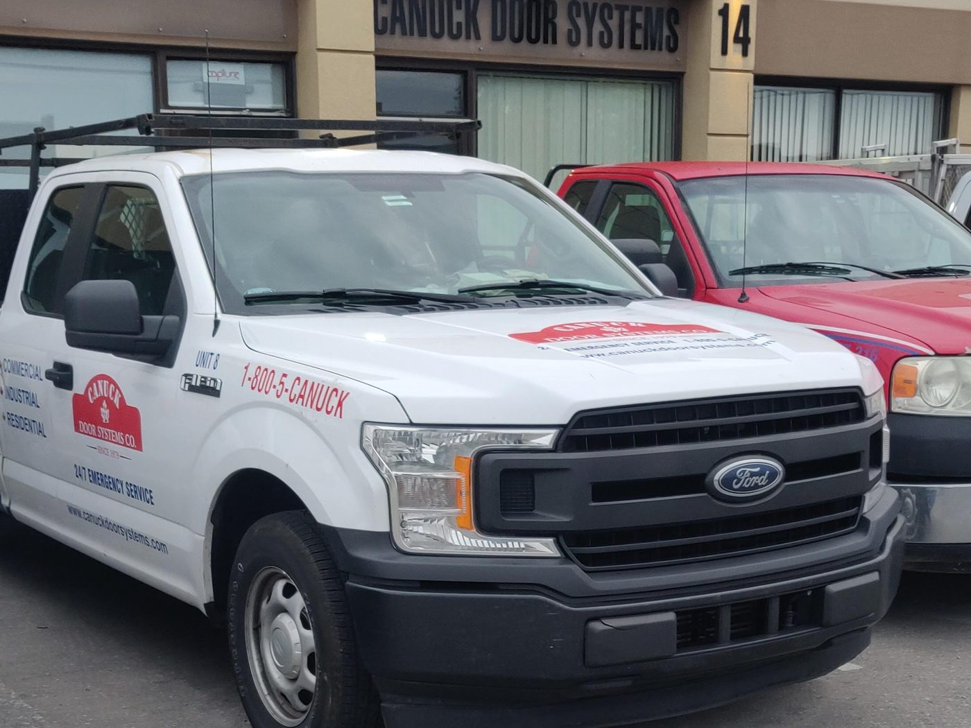 A Canuck Door Systems service truck with a roof rack, parked outside the company's shop at unit 14, 1645 Bonhill Road, Mississauga, under the sign on the building.