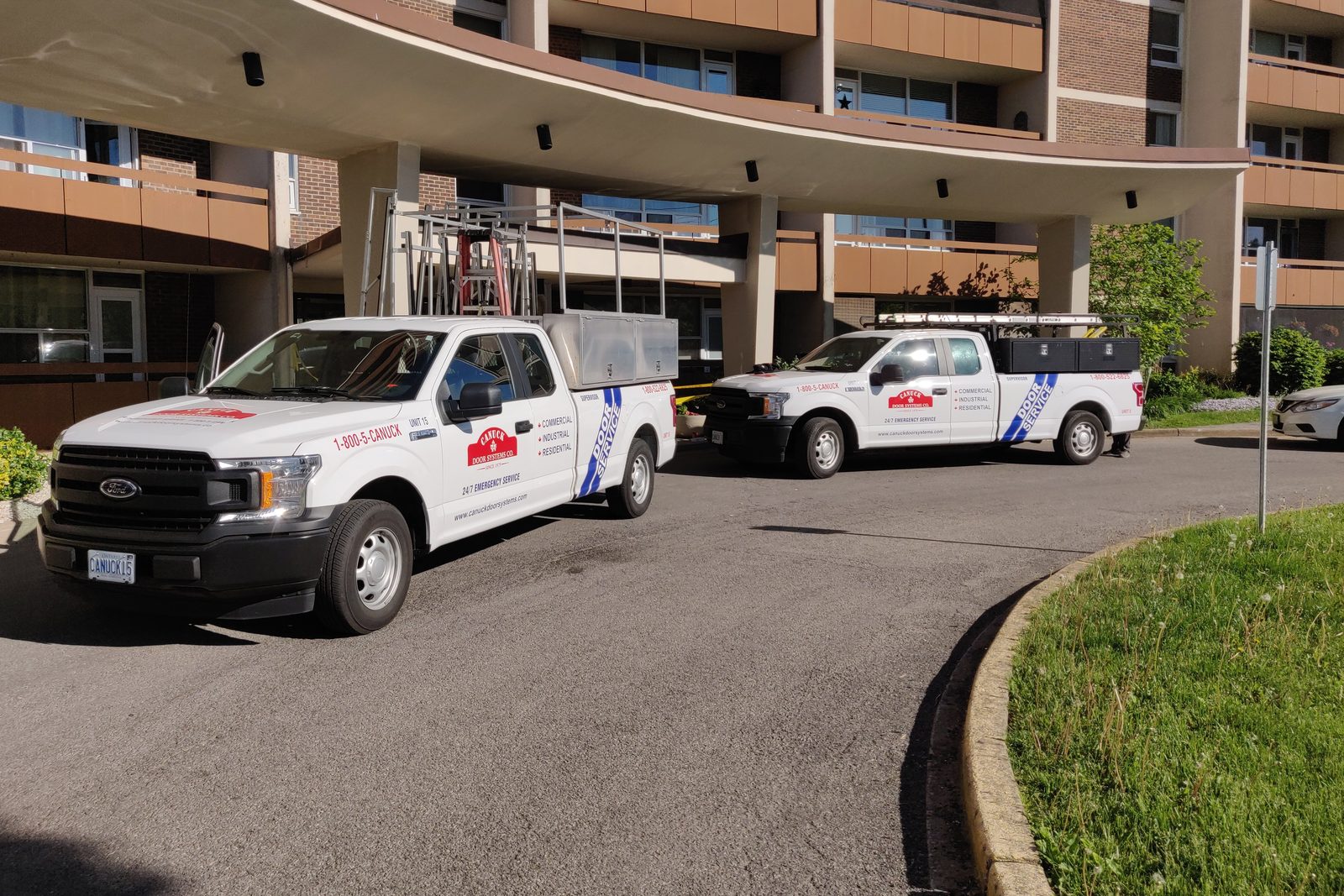 Two Canuck Door Systems service trucks carrying glass racks, parked under the entrance canopy of an apartment building on a service call
