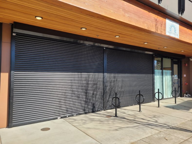 Two black roll-up security shutters closed over a row of storefront windows under a wood-slat soffit, with the shop's glass entrance beside them and bike racks on the sidewalk