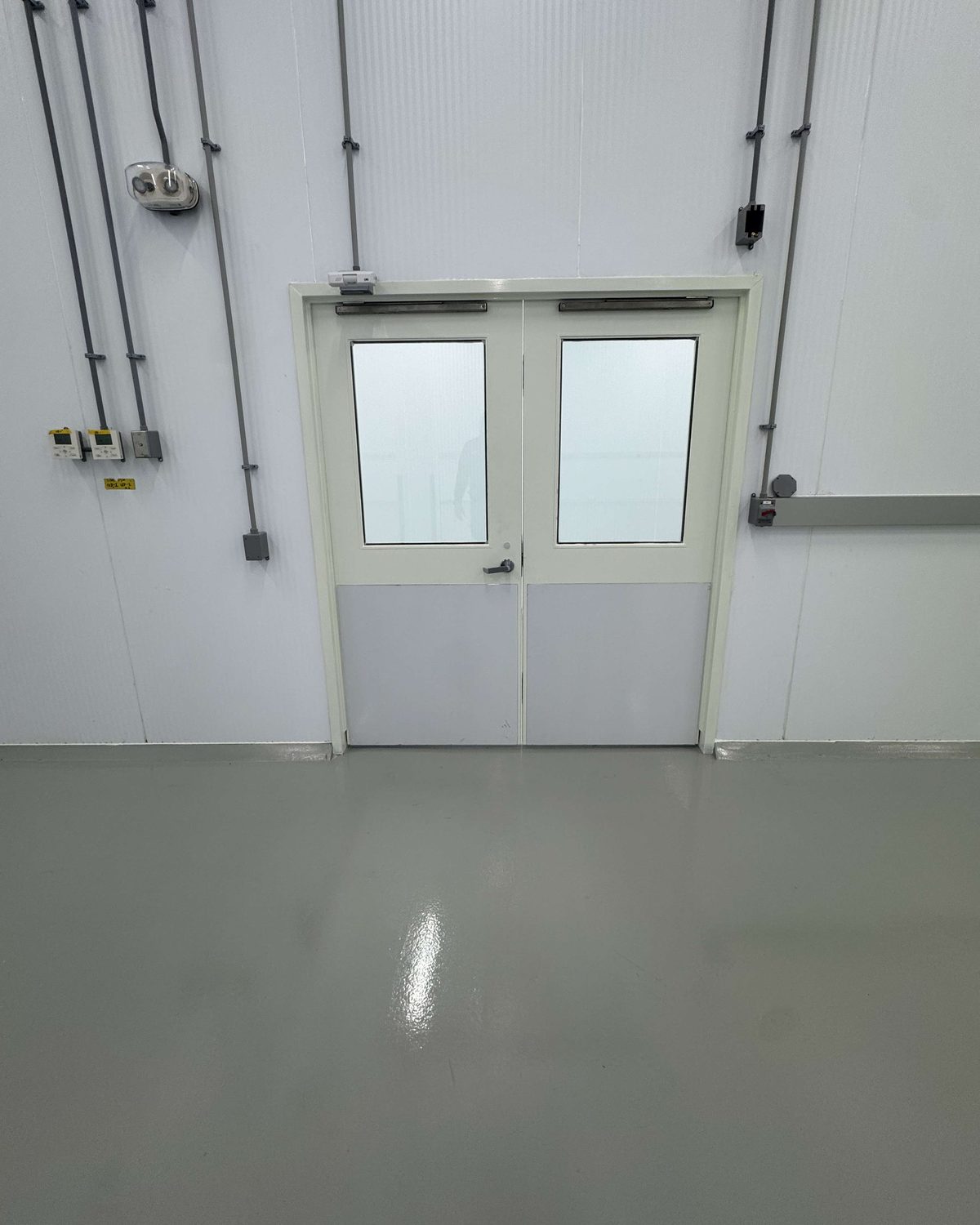 A pair of white hollow metal doors with vision lites at the end of a white-walled corridor inside Curaleaf's facility in Brampton, with conduit on the wall above
