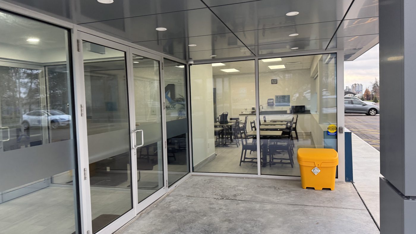 The glazed entrance vestibule of the Metelix expansion in Brampton under its canopy, with a pair of aluminum-framed glass doors and a glazed wall looking into a lunchroom