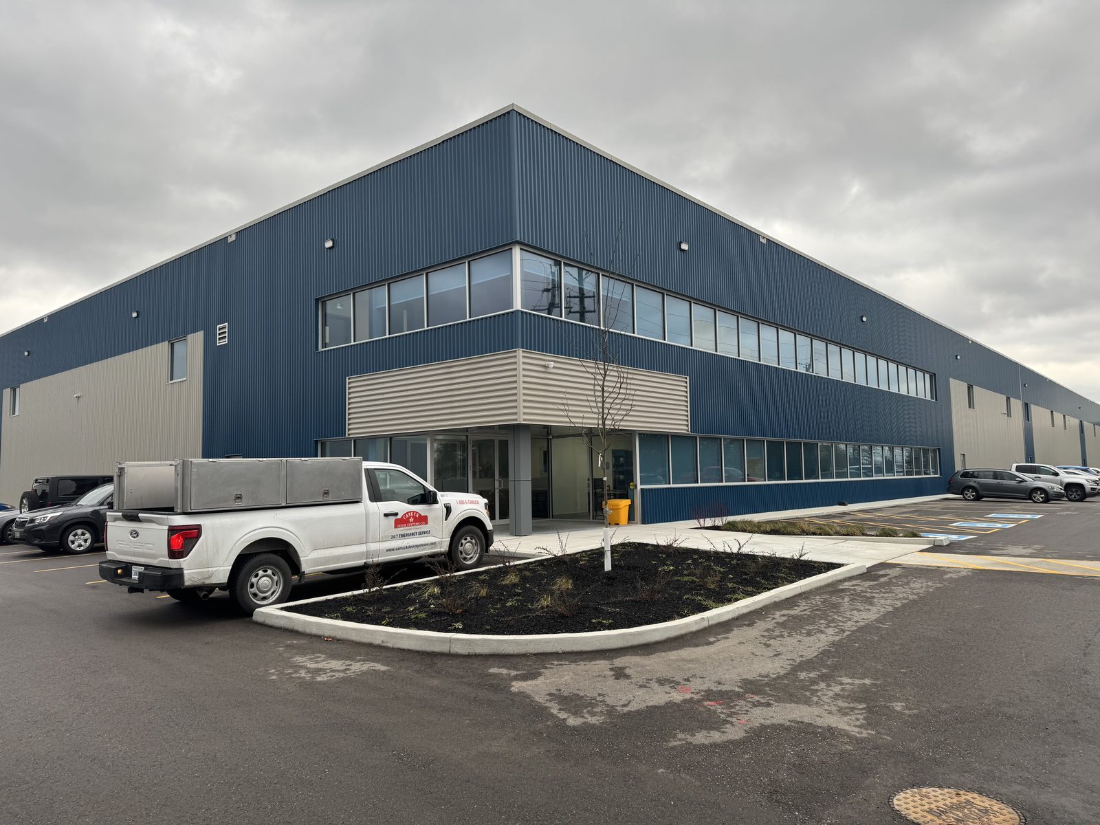 The finished two-storey expansion at 95 Van Kirk Drive, Brampton, with blue metal cladding, a continuous band of aluminum-framed windows on the upper floor and a glazed entrance under a canopy, a Canuck Door Systems service truck parked in front