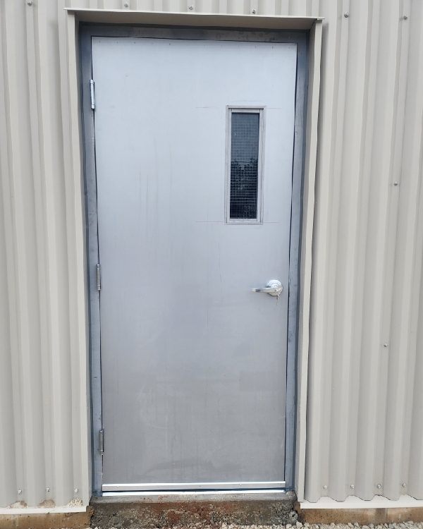 A grey hollow metal door with a narrow wired-glass vision lite and lever handle, set in a metal frame in beige corrugated cladding