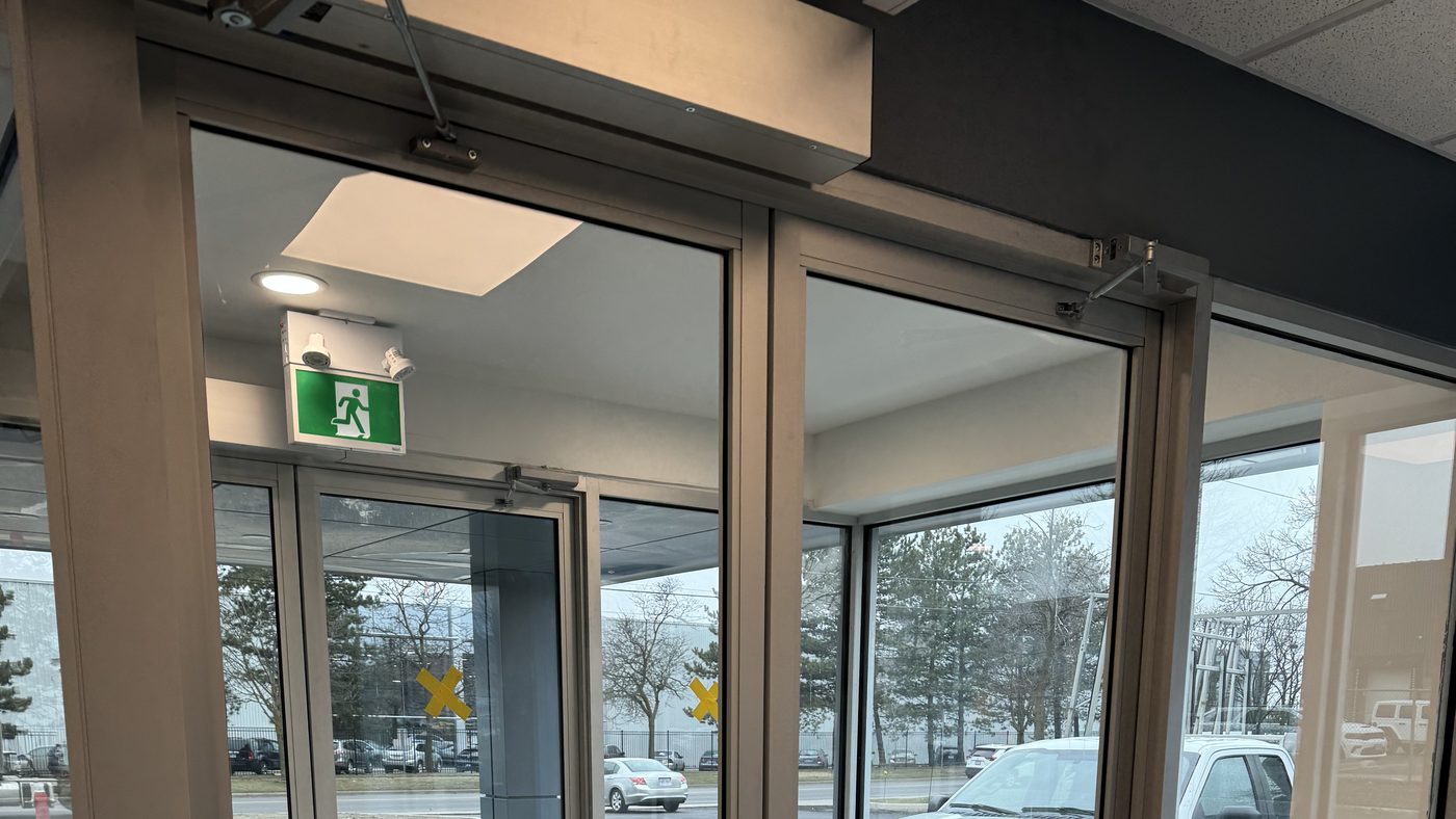 Inside the vestibule of the Metelix building in Brampton: a pair of aluminum-framed glass doors, an automatic operator housing with its arm above the left leaf, a closer on the right leaf and an exit sign beyond