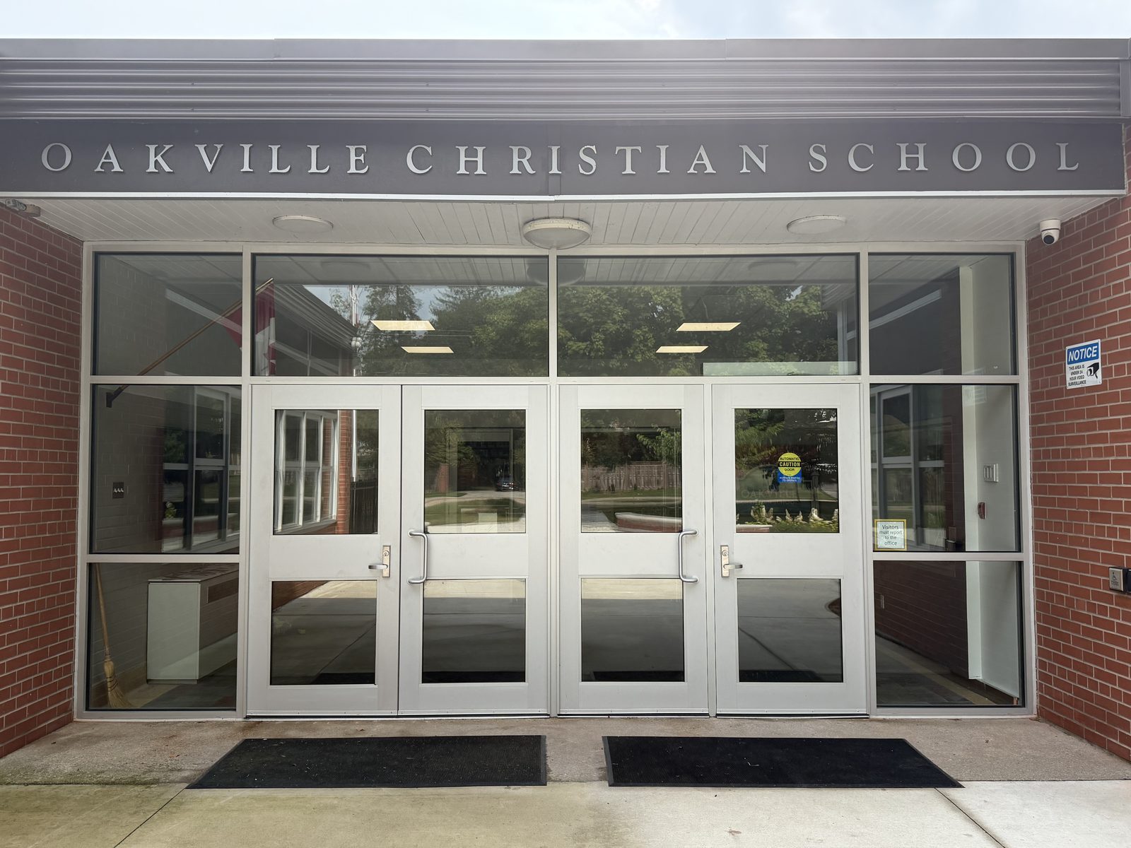 Four aluminum entrance door leaves in one glazed opening at Oakville Christian School — two double-door pairs either side of a mullion, glazed transoms above and sidelites to each side, entrance mats on the concrete, under a wall sign reading Oakville Christian School