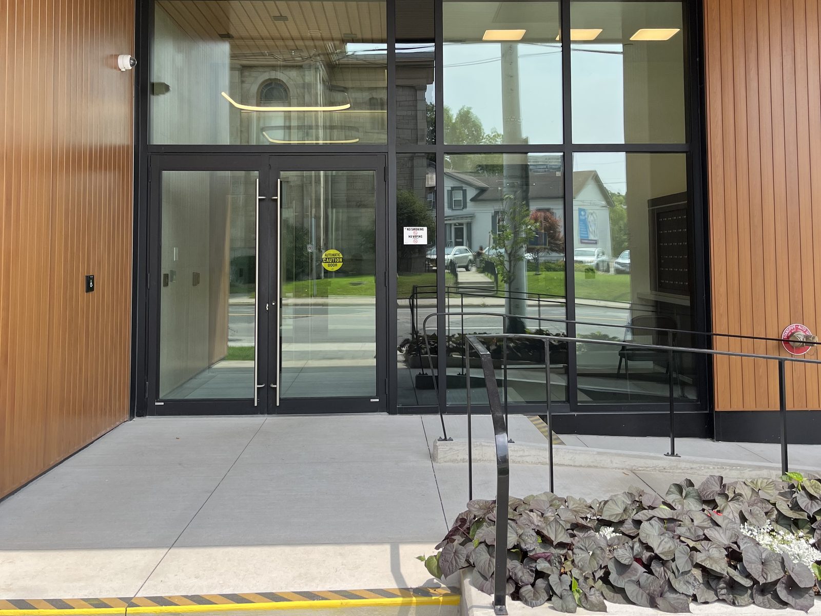 A glazed entrance of dark-framed curtain wall and double doors under a wood-slat soffit on Main Street in Dundas, with a ramp and planter in front, installed by Canuck Door Systems