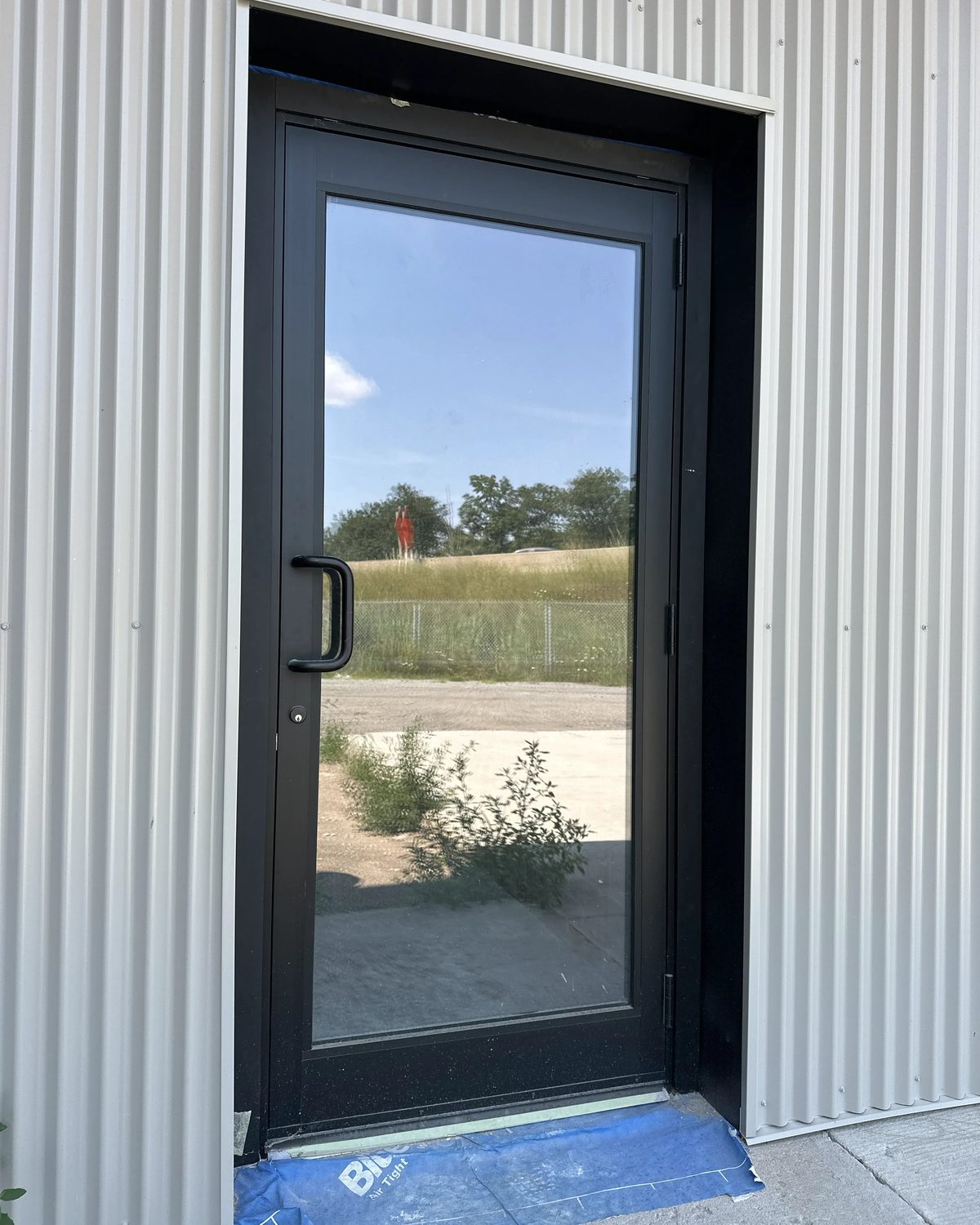 A black narrow-stile aluminum door with a full-height glass lite, set into the white corrugated steel wall of the industrial building at the same Hamilton site