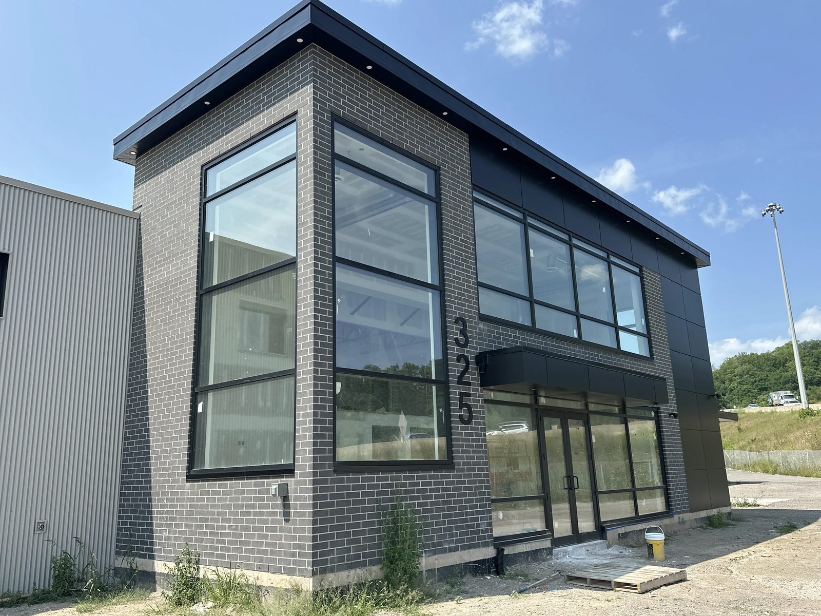 The finished two-storey office front on an industrial building in Hamilton — two-storey black-framed aluminum windows meeting at a dark brick corner pier, a ribbon of second-floor glazing, and a glazed storefront entrance under a black canopy, installed by Canuck Door Systems