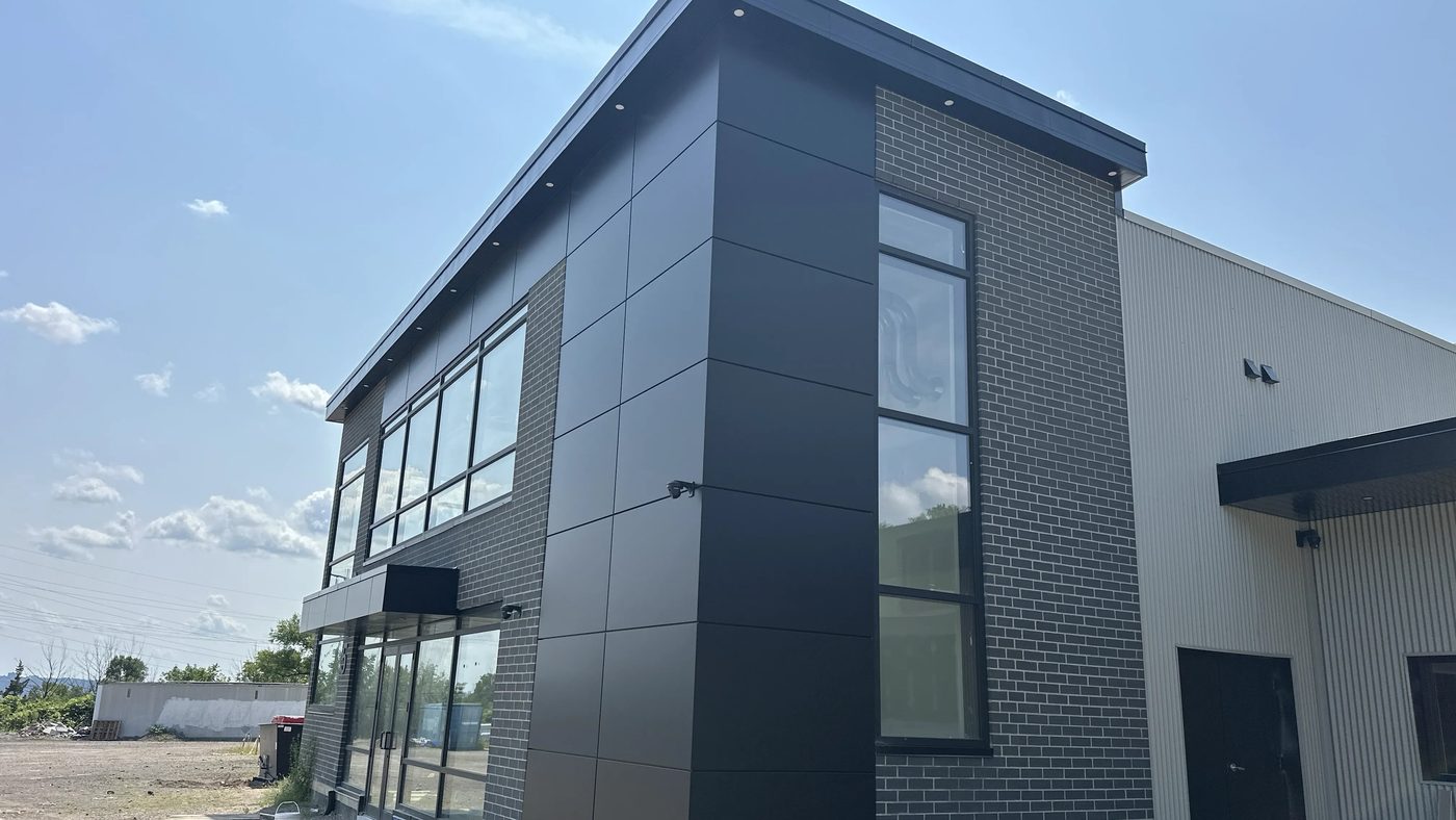 The new two-storey glazed office block seen from the side, where its dark brick and black metal panel meet the white corrugated steel of the existing industrial building in Hamilton