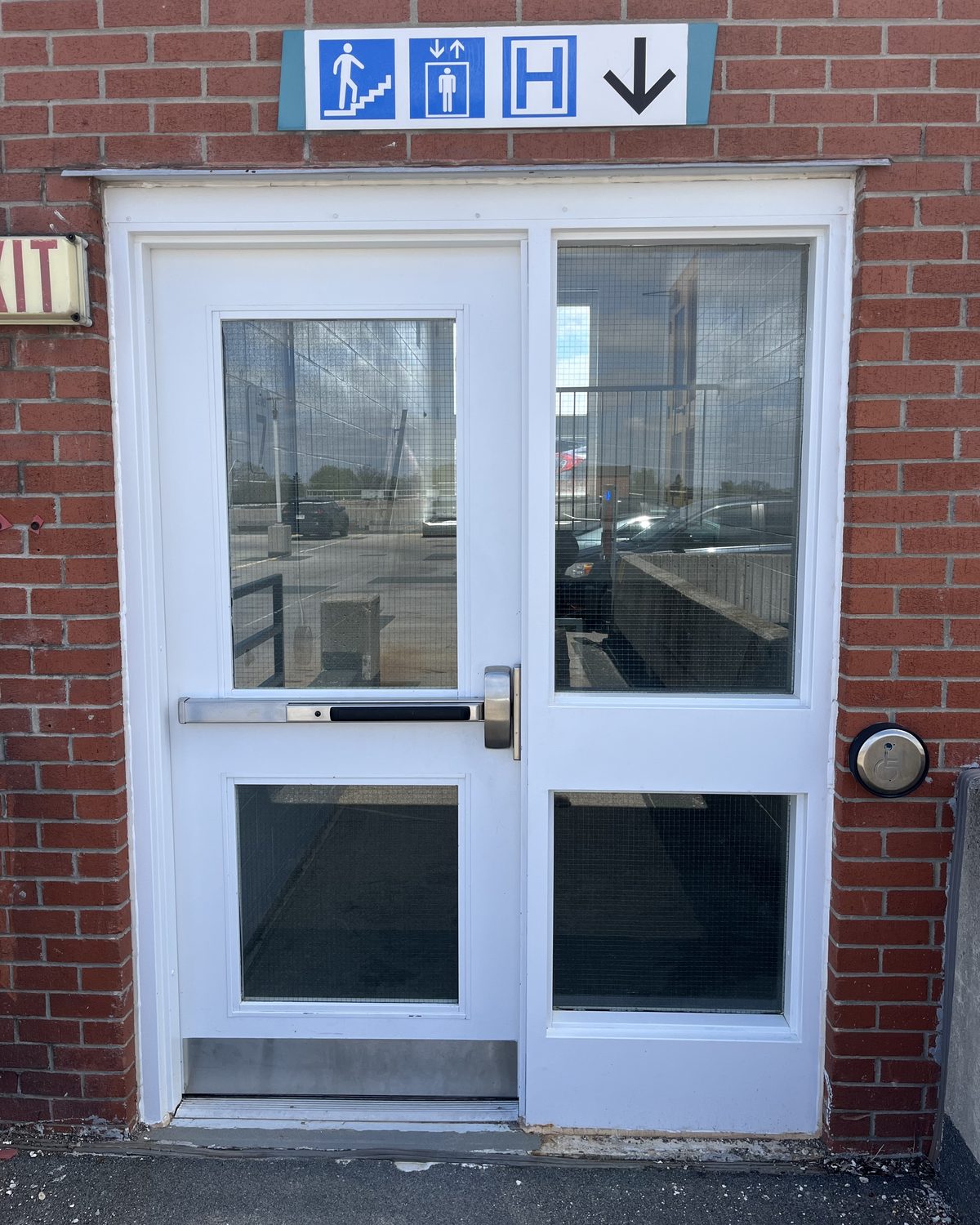 A pair of white hollow metal doors with vision lites and a panic bar in a red brick wall of the Grand River Hospital parking garage in Kitchener, under an accessibility sign