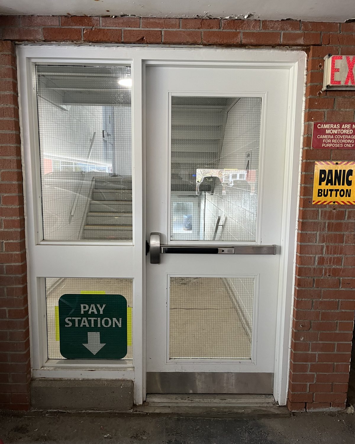 A white hollow metal door with a vision lite and a panic bar, with a glazed sidelite showing the stairs behind, in the Grand River Hospital parking garage in Kitchener