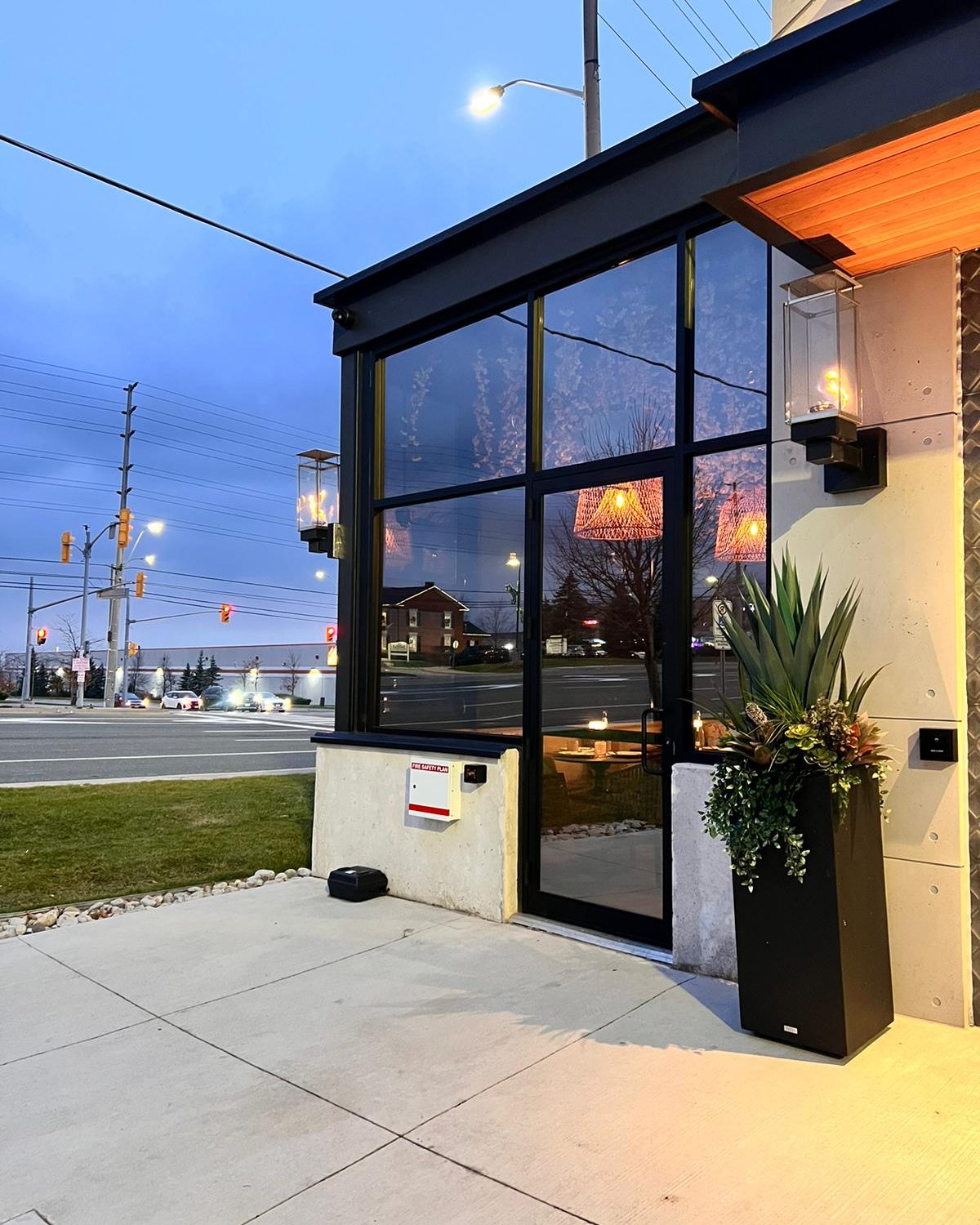 The glazed corner of the Mishree restaurant in Mississauga at dusk, floor-to-ceiling glass in dark aluminum framing beside a planter