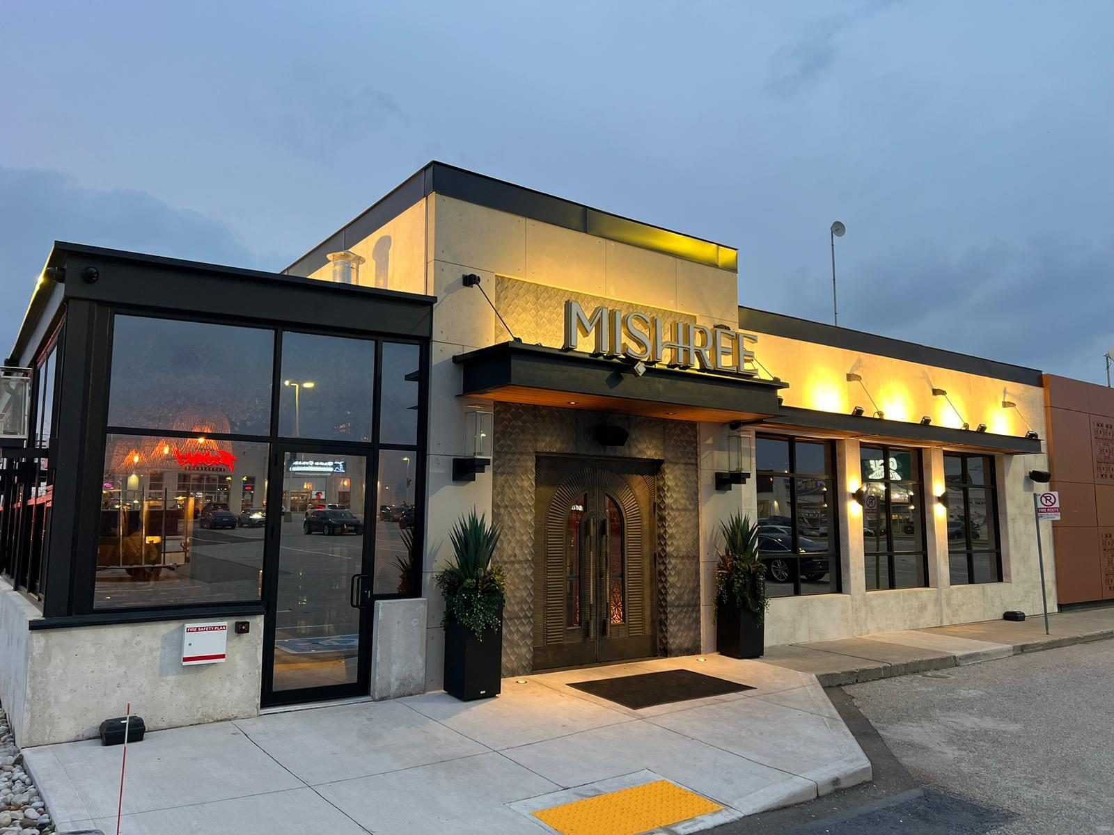 The front of the Mishree restaurant in Mississauga at dusk, with dark aluminum storefront glazing, a glazed corner and the entrance lit beneath the restaurant's sign, installed by Canuck Door Systems