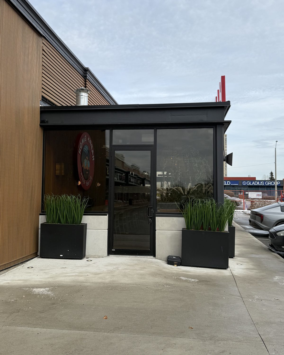 A glazed entrance set into the dark aluminum front at the corner of the Happy Singh restaurant in Mississauga, with planters in front