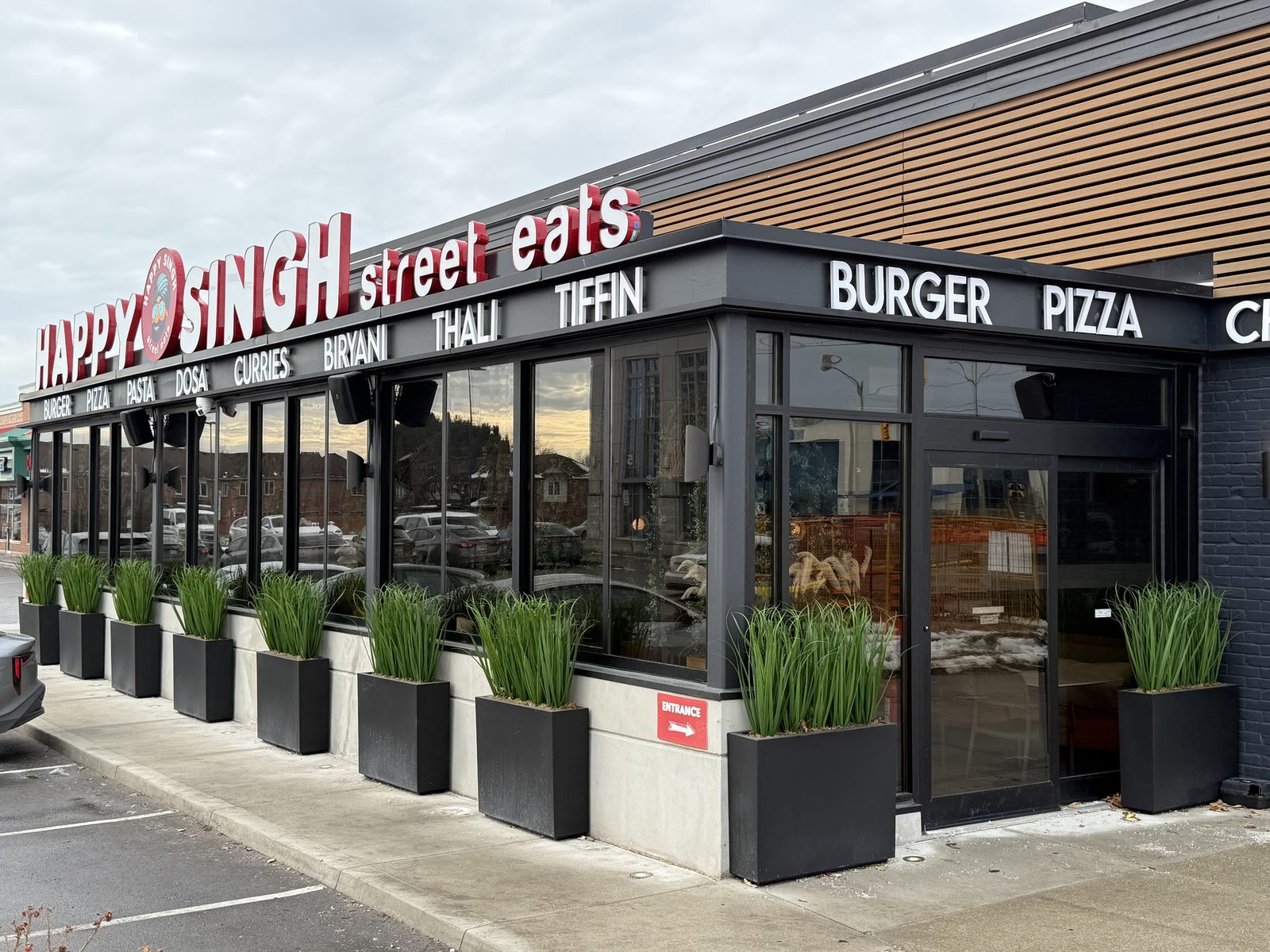 The glazed front of the Happy Singh restaurant in Mississauga: a long run of dark aluminum storefront glazing and doors under the restaurant's sign, with planters along the sidewalk, installed by Canuck Door Systems