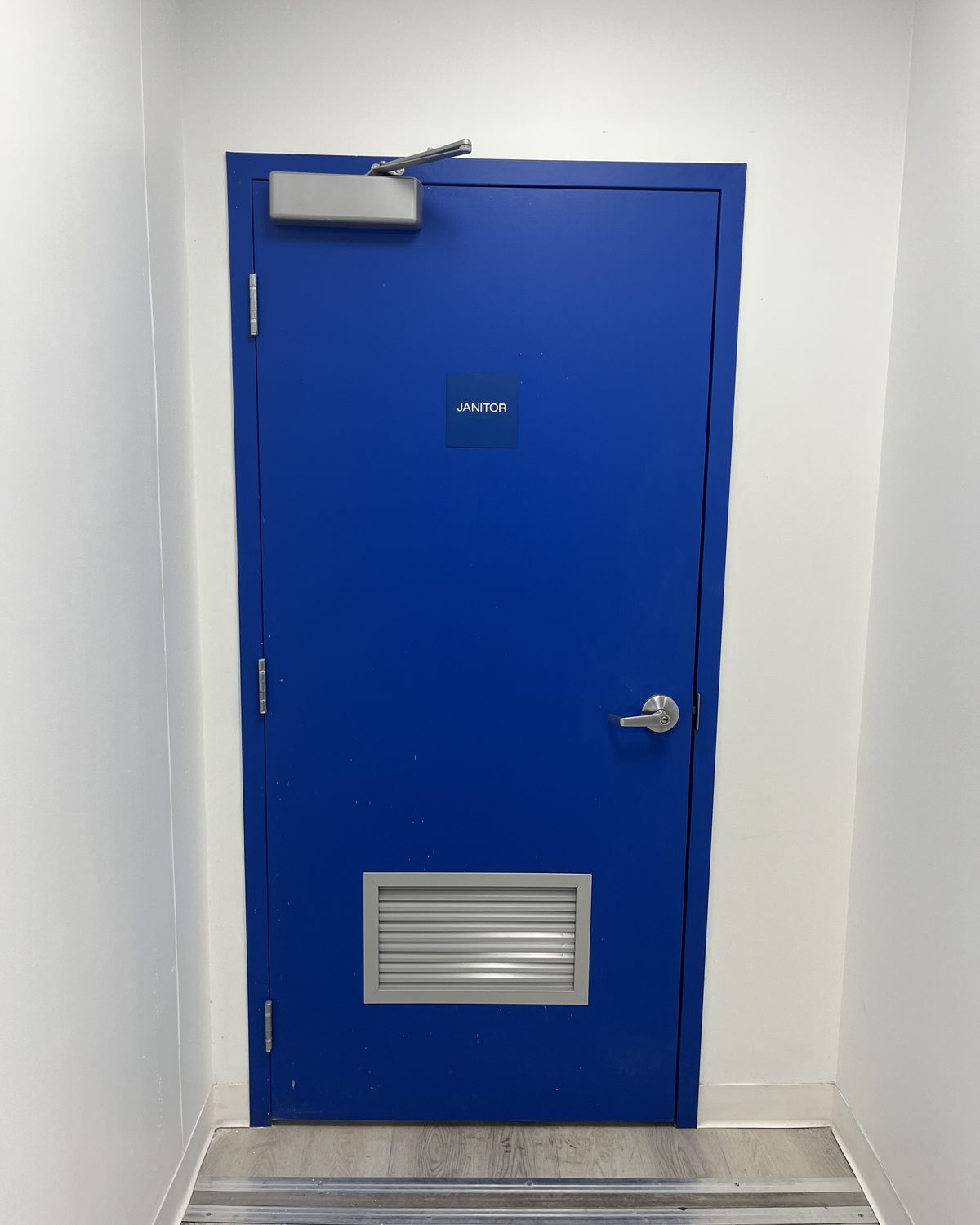 A blue hollow metal door with a louver in its lower panel and a closer above, in a white corridor at a Toys “R” Us store in North York