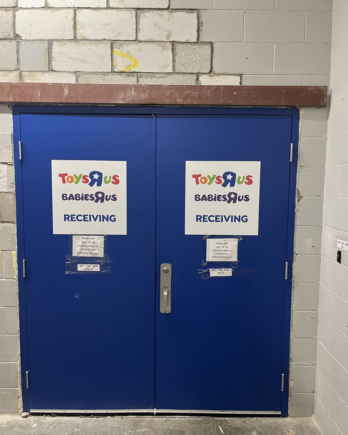 A pair of blue hollow metal receiving doors in a block wall at a Toys “R” Us store in North York, with store signs taped to each leaf