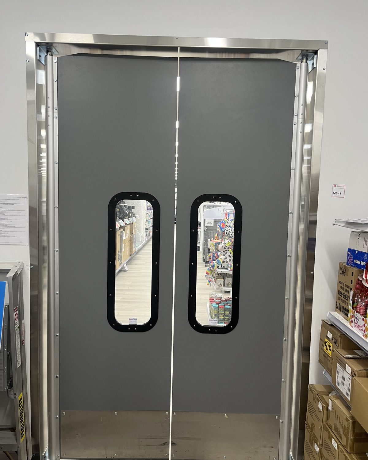 Grey double traffic doors with round-cornered vision windows in a stainless steel frame, in the back of a Toys “R” Us store in North York