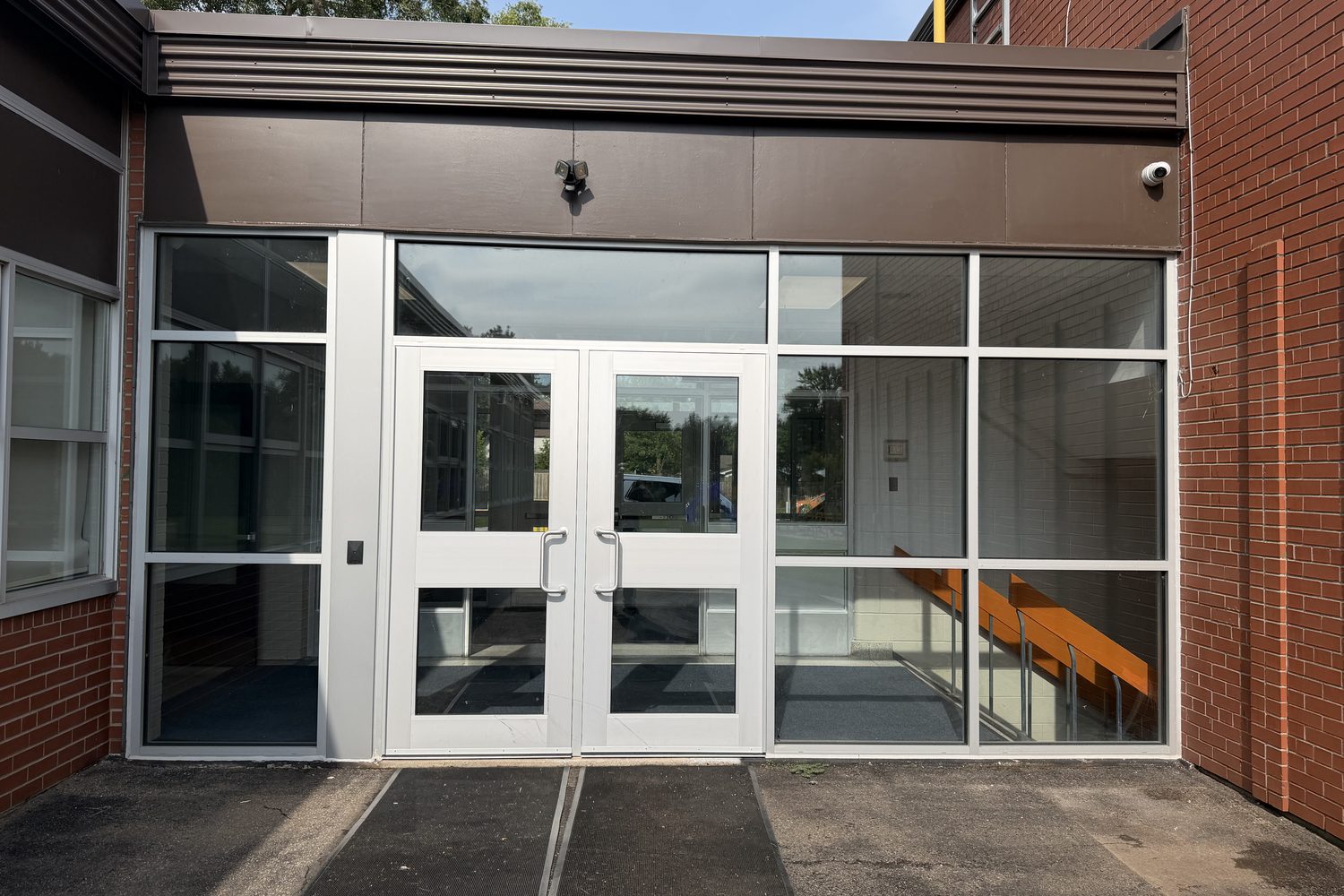 An aluminum double-door entrance in a brick elevation at Oakville Christian School, seen under the building's canopy — a glazed transom above the doors, a solid aluminum infill panel to one side and glazed sidelites to the other