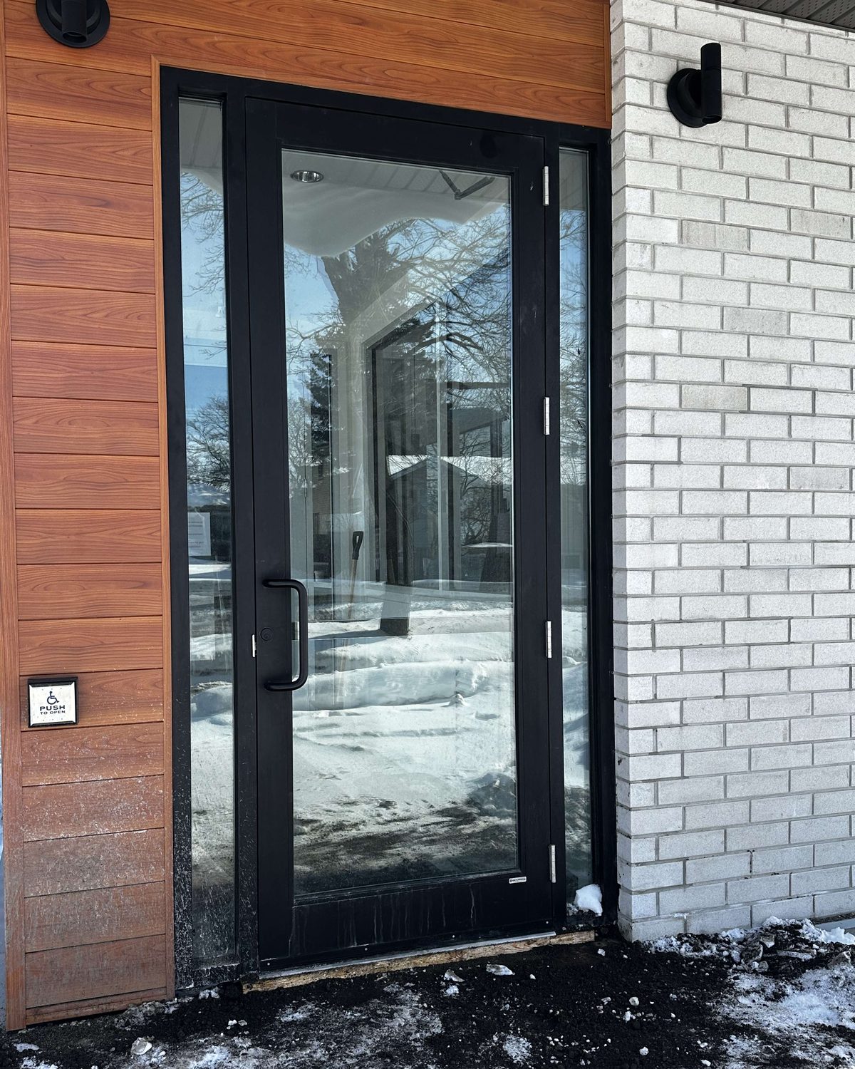 The glazed aluminum entrance door of Shelburne Dental in Shelburne, set between wood-look cladding and white brick, with an accessible push button beside it