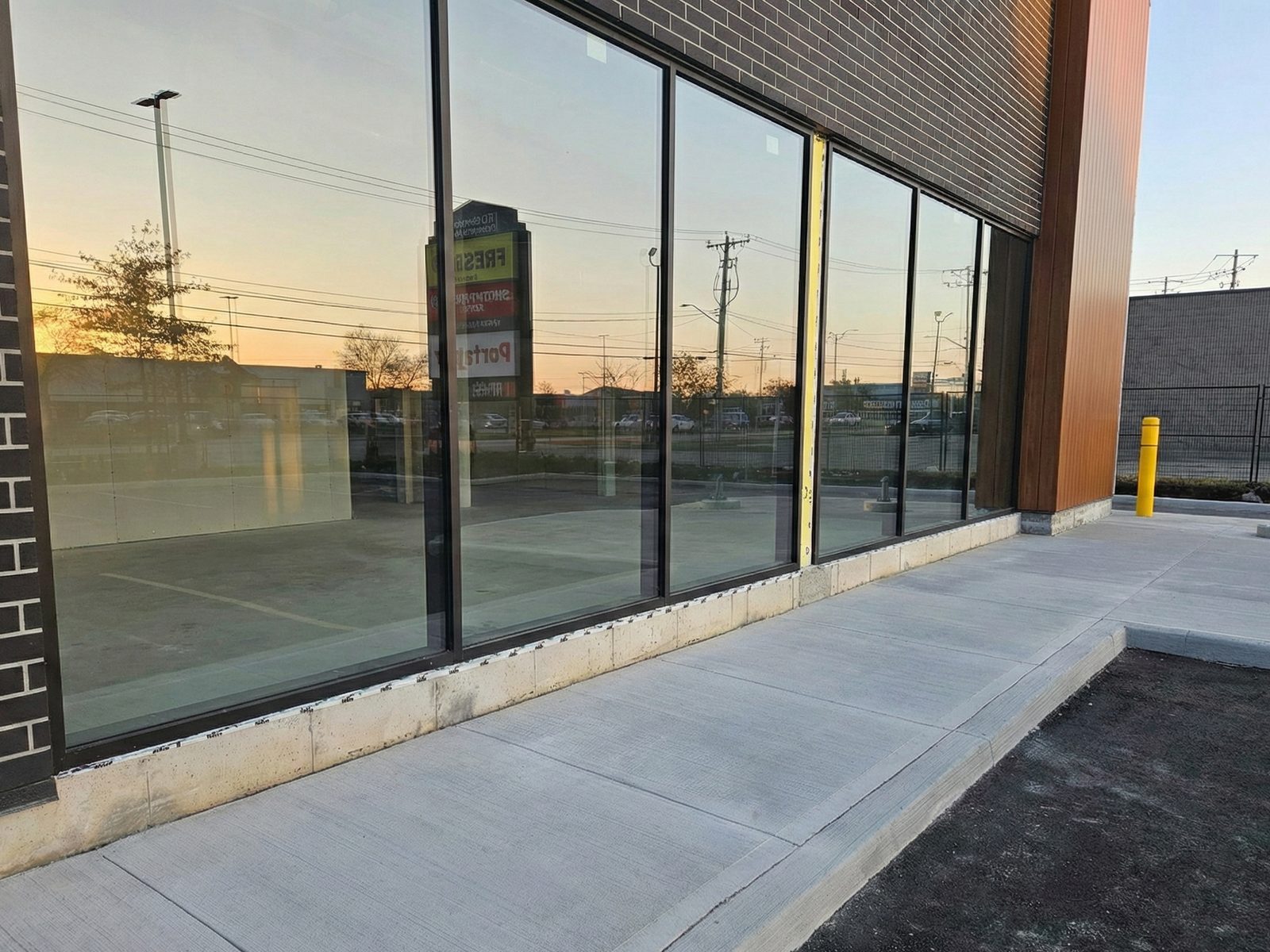 A new run of dark aluminum storefront windows along the base of a brick-clad plaza building at 286 Bunting Road in St. Catharines, with a new sidewalk in front, installed by Canuck Door Systems