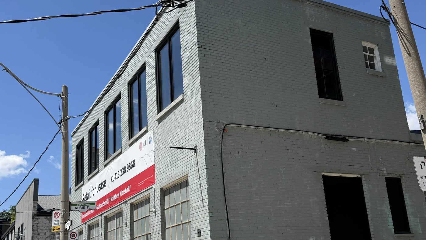 The street corner of a two-storey painted brick building in Toronto with new black-framed second-floor windows and a Retail for Lease banner, with a condominium tower behind, installed by Canuck Door Systems